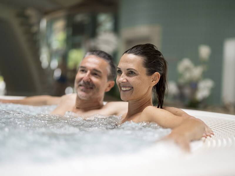 Zwei Personen entspannen in einem Whirlpool mit sprudelndem Wasser in einem Innenbereich.