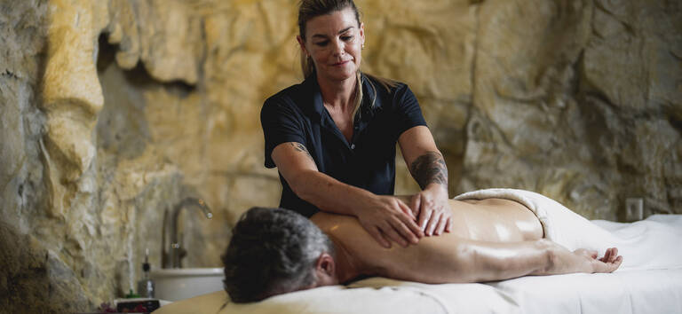 Masseurin massiert Rücken eines liegenden Mannes in einem Raum mit Felswand im Hintergrund.