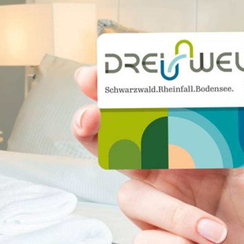 Hand hält eine DREI WELTEN CARD mit grün-blauem Design vor einem Hotelzimmer mit Bett, Lampe und weißen Handtüchern.
