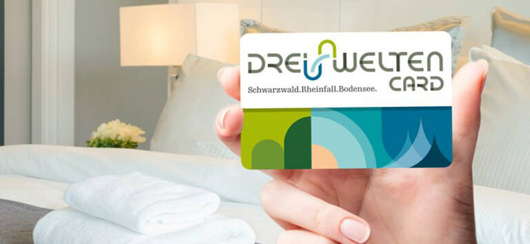 Hand hält eine DREI WELTEN CARD mit grün-blauem Design vor einem Hotelzimmer mit Bett, Lampe und weißen Handtüchern.