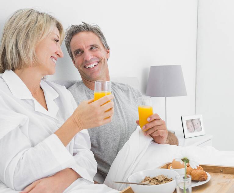 Paar im Bett mit weißer Bettwäsche, das Orangensaft trinkt, auf einem Tablett mit Frühstücksflocken, Croissant und Glas mit Blume.