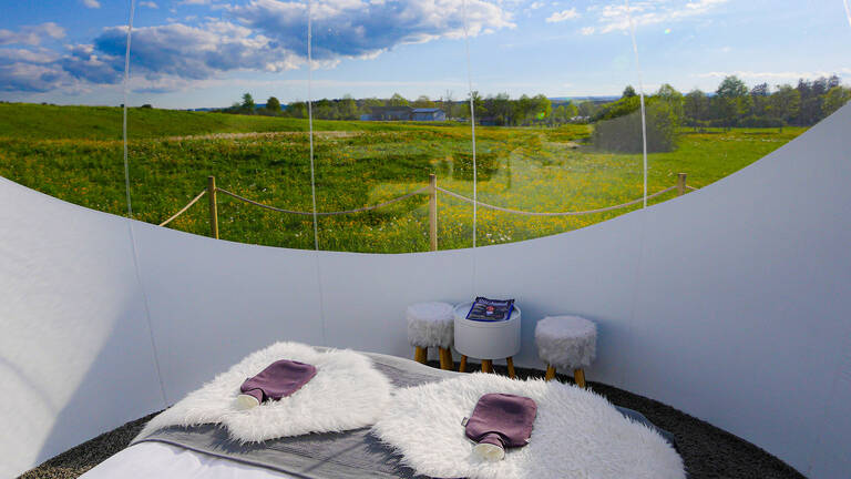 Innenansicht eines transparenten Bubble-Zelts mit zwei Liegen, weißen Fellauflagen, zwei Handtüchern und einem kleinen Tisch vor einer grünen Wiese unter blauem Himmel.