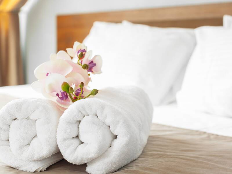 Zwei aufgerollte weiße Handtücher mit Orchideenblüten auf einem Bett mit weißen Kissen