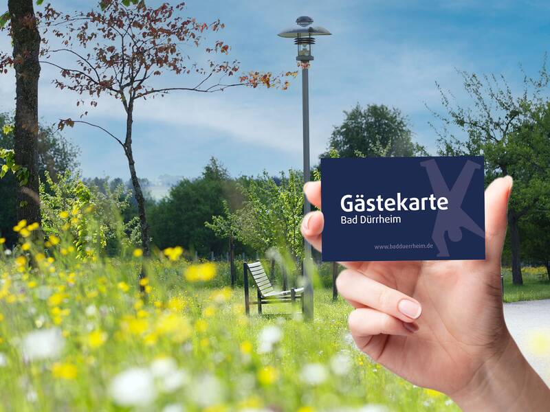 Hand hält Gästekarte Bad Dürrheim vor einer Parklandschaft mit Blumen und Bänken