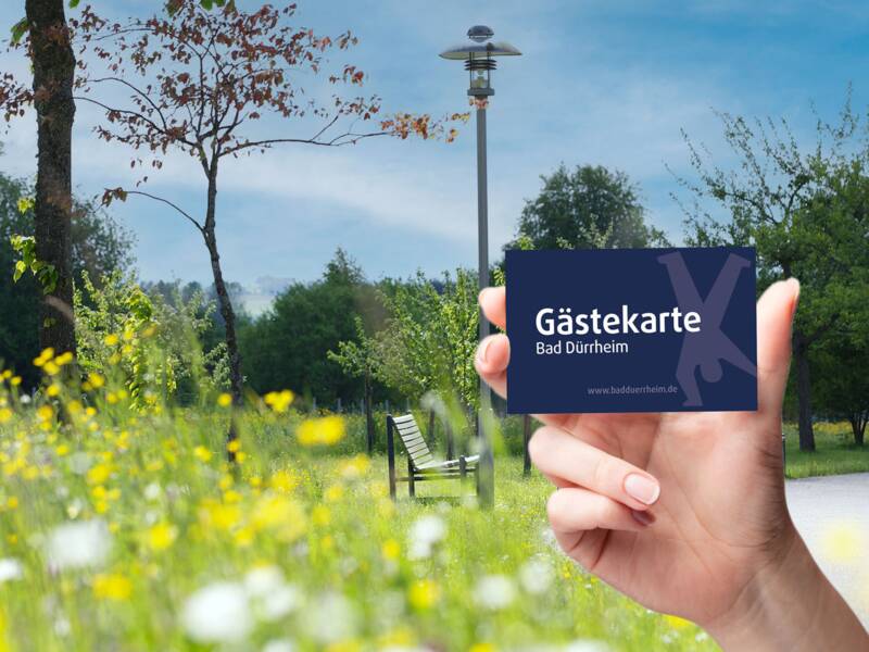 Hand hält Gästekarte Bad Dürrheim vor einer Parklandschaft mit Blumen und Bänken