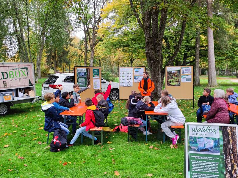 Kinder sitzen an Tischen im Freien auf einer Wiese, ein Erwachsener steht vor mehreren Infotafeln unter Bäumen, im Hintergrund ein Anhänger mit der Aufschrift 'DIE WALDBOX'