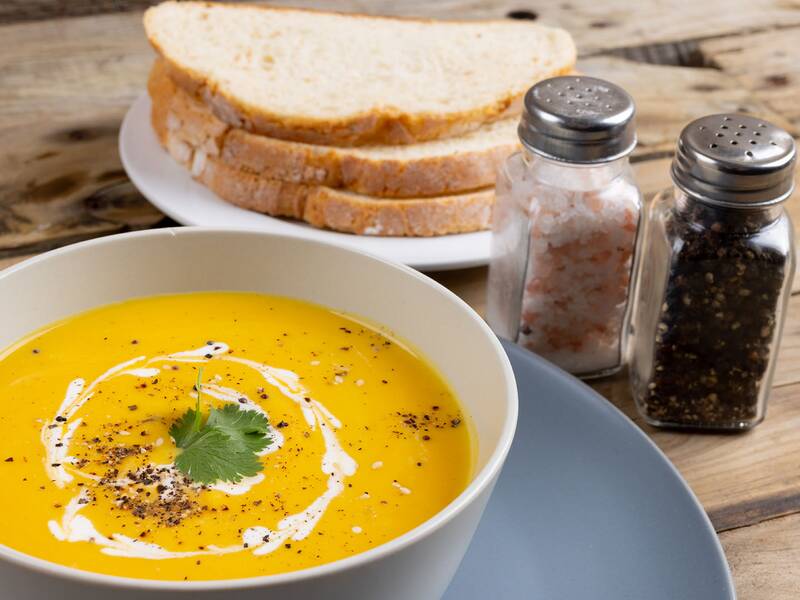 Schale mit cremiger Suppe, garniert mit einem Blatt Koriander und einem Sahnewirbel, daneben Salz- und Pfefferstreuer sowie mehrere Scheiben Brot auf einem Holztisch