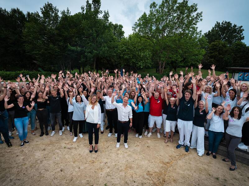 Große Gruppe von Menschen steht im Freien auf sandigem Boden und hebt die Arme, umgeben von Bäumen und einem Holzsteg.