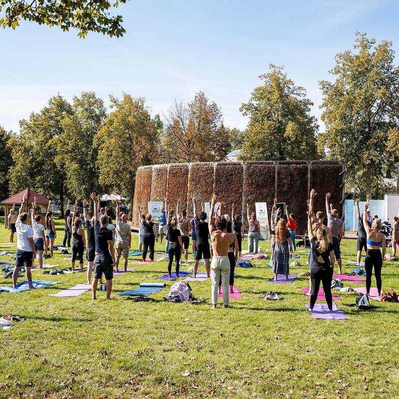 Mehrere Menschen stehen auf Yogamatten auf einer Wiese und heben die Arme nach oben, im Hintergrund Bäume und eine Nachbildung eines Gradierwerks.