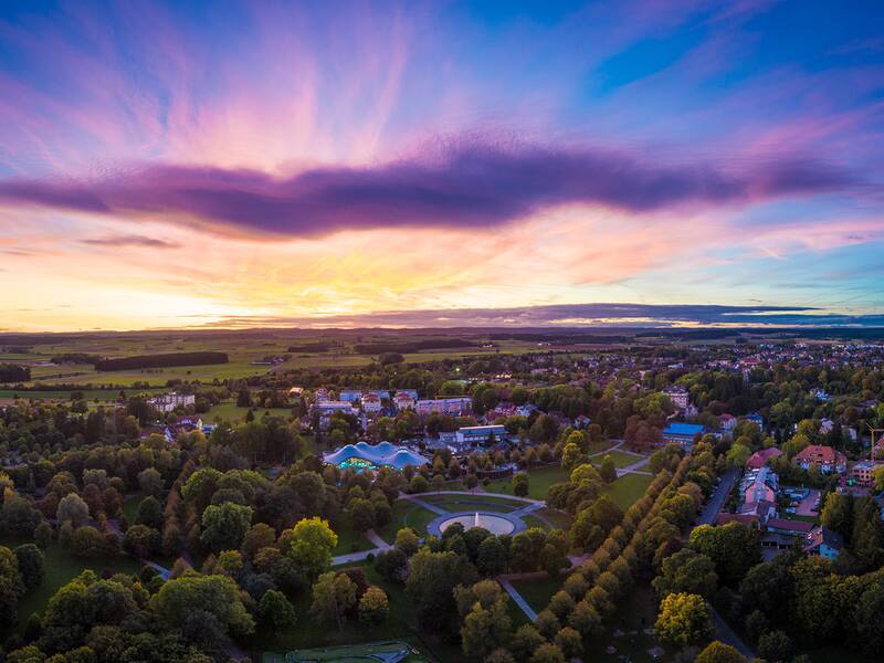 Luftaufnahme eines Parks mit Wegen, Bäumen und Gebäuden bei farbigem Abendhimmel mit Wolken.