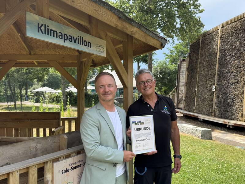 Zwei Männer stehen vor einem Haus und halten eine Urkunde mit der Aufschrift 'Premium Class' und eine weitere bedruckte Karte mit dem Logo 'Heilklima Wandern'