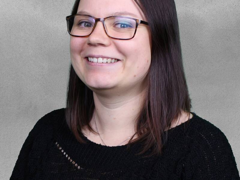 Frau mit schulterlangem, glattem Haar und schwarzem Pullover vor grauem Hintergrund.