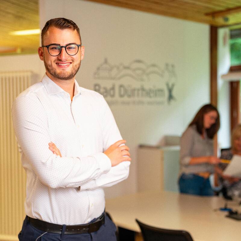 Ein Mann mit Brille steht aufrecht mit den Armen verschränkt da und lächelt in die Kamera. Er trägt ein weißes Hemd und eine blaue Stoffhose. Im Hintergrund sind zwei Frauen zu erkennen, die sich zusammen an einem Schreibtisch über Unterlagen beugen.