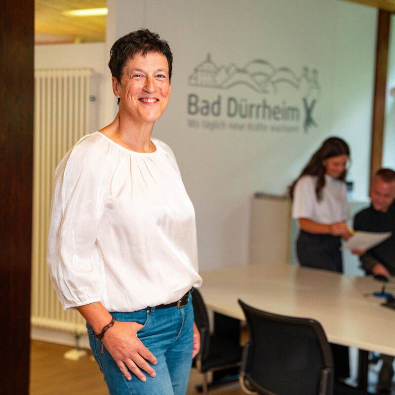 Frau mit kurzen, dunklen Haaren steht aufrecht und locker da und lächelt in die Kamera. Sie trägt eine weiße Bluse und Jeans-Hose. Im Hintergrund sind eine weitere Frau und ein Mann zu erkennen, die sich zusammen an einem Schreibtisch über Unterlagen beugen.
