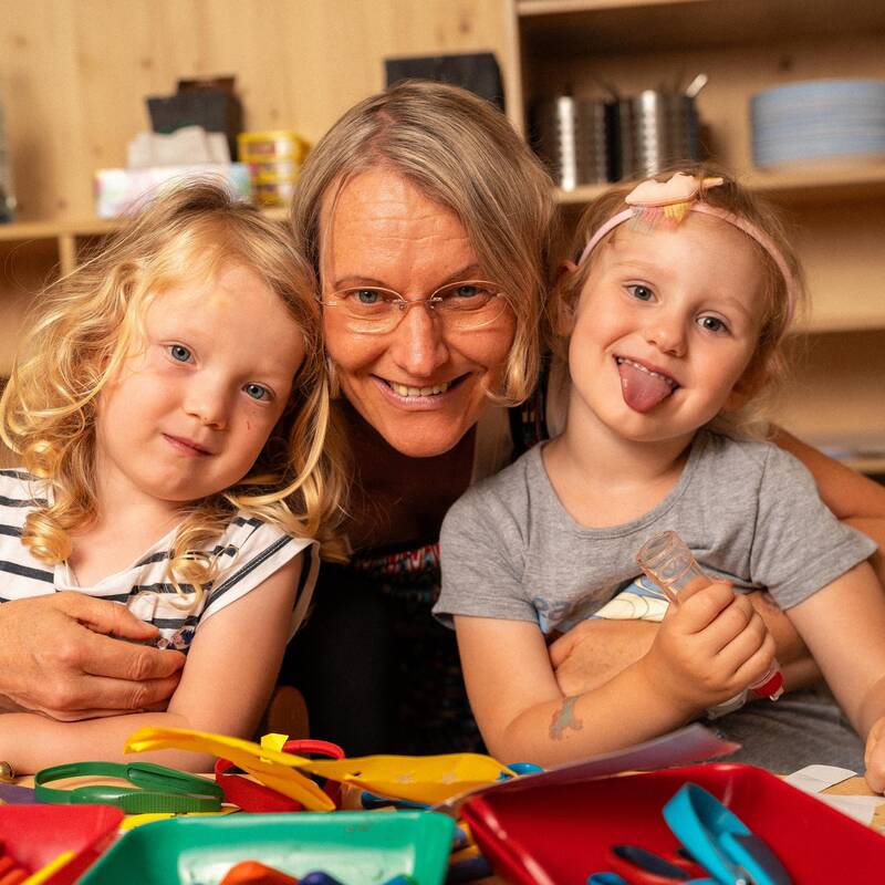 Eine Frau hält zwei Kinder in den Armen und schaut lächelnd in die Kamera. Die zwei Mädchen links und rechts schauen in die Kamera, das rechte Mädchen streckt die Zunge raus. 