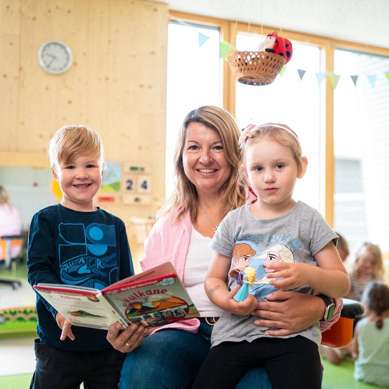 Eine Frau mit schulterlangen blonden Haaren kniet auf dem Boden und hat ein Buch in der rechten Hand und schaut lächelnd in die Kamera. Die linke Hand liegt um ein Mädchen, was links neben dran steht schüchtern in die Kamera schaut. Rechts steht noch ein Junge, welcher fröhlich in die Kamera lächelt. 