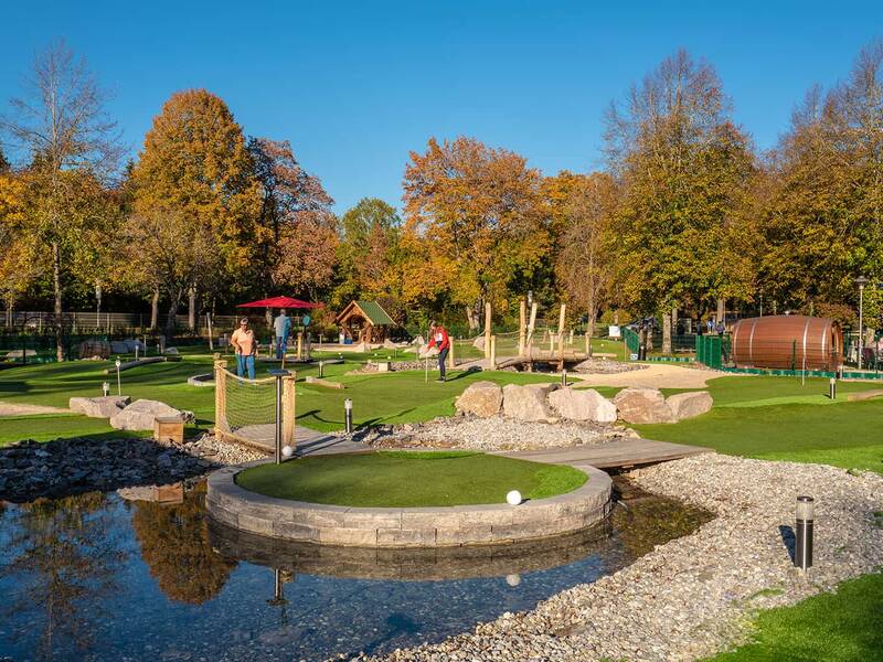 Adventure-Golf-Anlage mit mehreren Bahnen, Steinen und Wasserhindernissen, umgeben von herbstlich gefärbten Bäumen und Menschen beim Spielen.