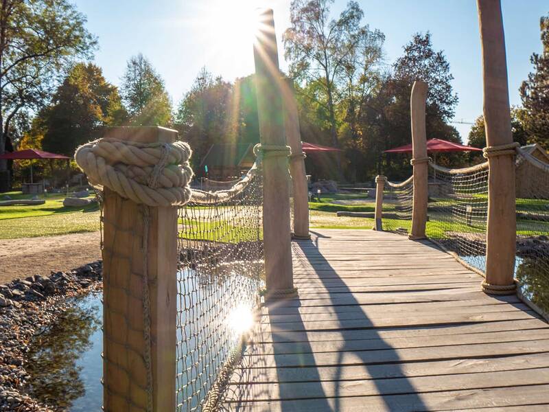 Holzbrücke mit dicken Seilen als Geländer über einem kleinen Teich, im Hintergrund Bäume und Sonnenschein.