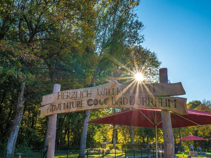 Holzschild mit der Aufschrift 'Herzlich Willkommen Adventure Golf Bad Dürrheim' vor Bäumen und Sonnenschein, roter Sonnenschirm im Hintergrund.
