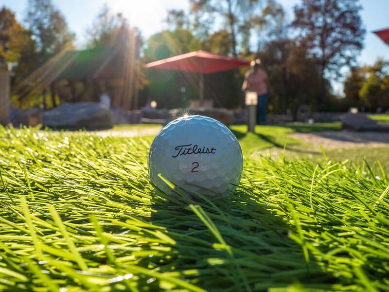 Weißer Titleist-Golfball mit roter Nummer 2 liegt auf grünem Kunstrasen, im Hintergrund unscharfe Person und Sonnenschirme.