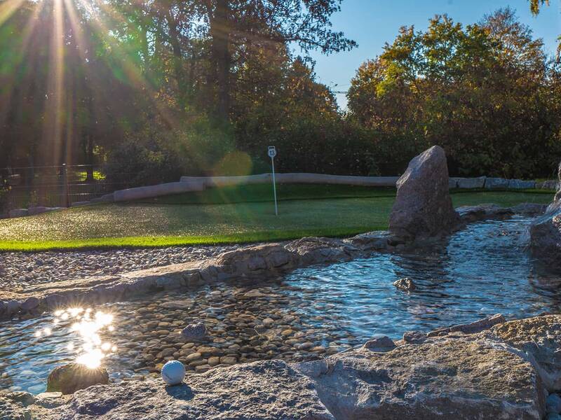 Adventure-Golf-Bahn mit grünem Kunstrasen, Wasserlauf mit Steinen und Golfball im Vordergrund, Sonne strahlt durch Bäume.