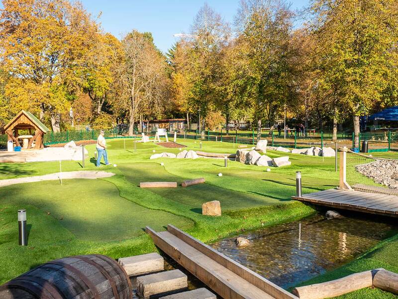 Adventure-Golf-Anlage mit mehreren Bahnen, Holzbrücke über Wasserlauf, herbstliche Bäume und zwei Personen auf dem Grün.