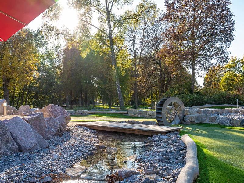 Wasserrad neben einem kleinen Bachlauf in einer Adventure-Golf-Anlage mit Steinen und grünem Kunstrasen.