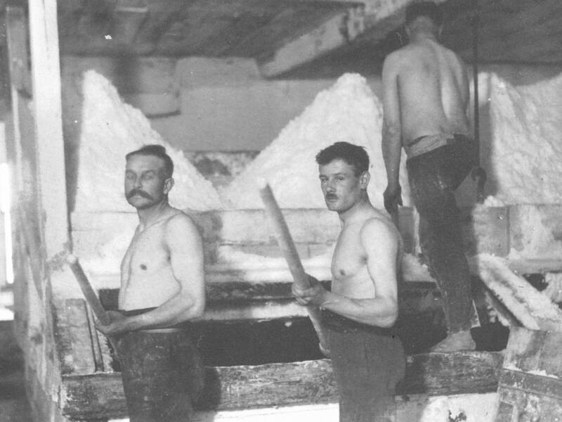 Drei Männer ohne Oberbekleidung arbeiten in einer Salzgewinnungsanlage, zwei halten lange Holzstangen, einer steht auf einer Holzrampe