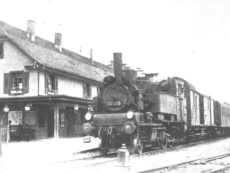 Schwarz-weiße Aufnahme einer Dampflokomotive vor einem Bahnhofsgebäude mit wartenden Personen