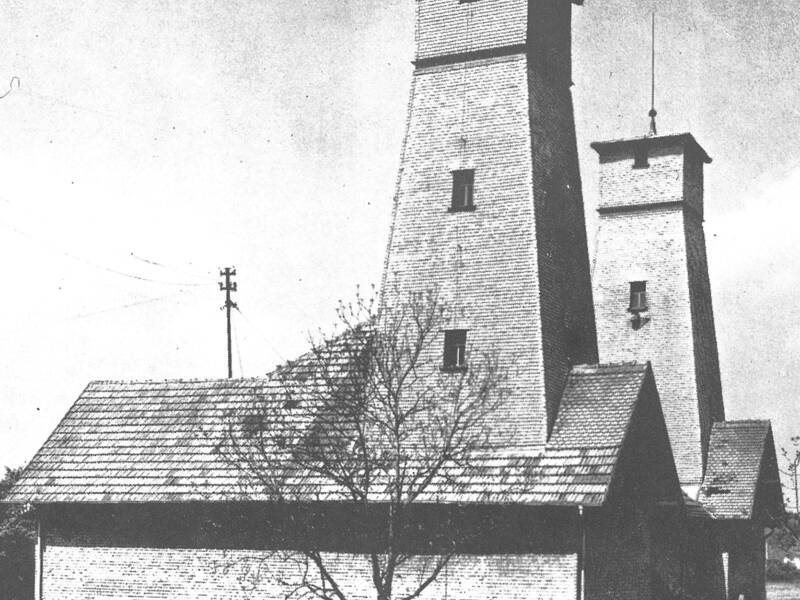 Schwarz-weiß-Fotografie eines Gebäudes mit zwei hohen, rechteckigen Türmen und einem niedrigeren Anbau mit Fenstern und einem Baum davor
