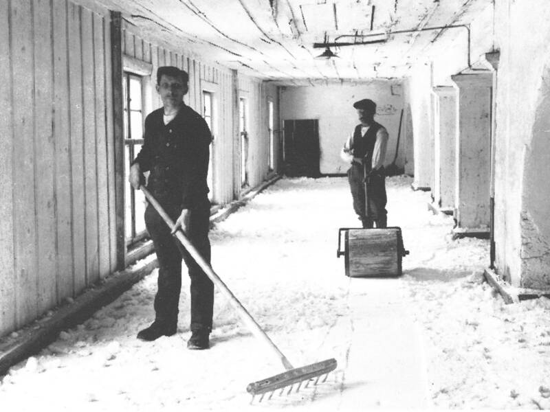 Zwei Männer in Arbeitskleidung in einem langen Raum, einer hält einen Rechen, der andere steht neben einem Kasten auf dem Boden