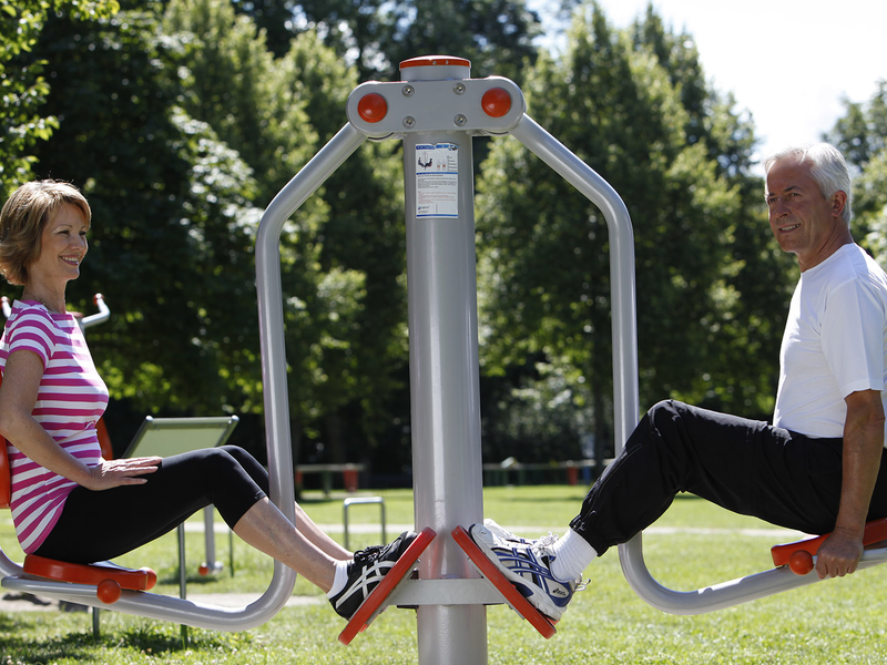Zwei Personen sitzen auf gegenüberliegenden Sitzen eines Fitnessgeräts im Freien, umgeben von Bäumen und Wiese.