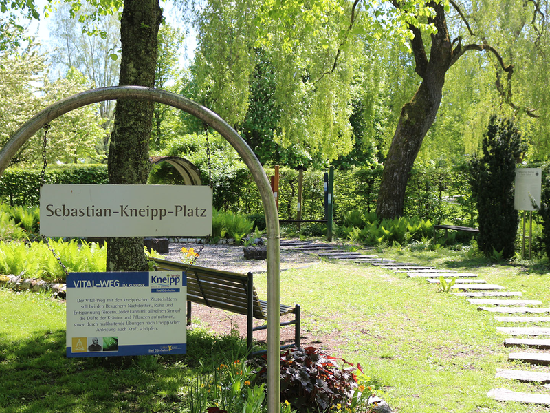 Bogenförmiges Schild mit der Aufschrift 'Sebastian-Kneipp-Platz' vor einer Parkbank und einem Weg mit Trittsteinen in einem grünen Park.