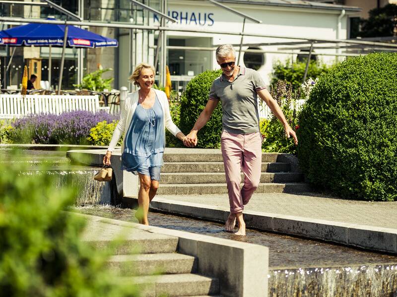 Paar hält Händchen und geht barfuß durch einen flachen Wasserlauf im Kurpark vor dem Kurhaus mit Sonnenschirmen und Büschen.