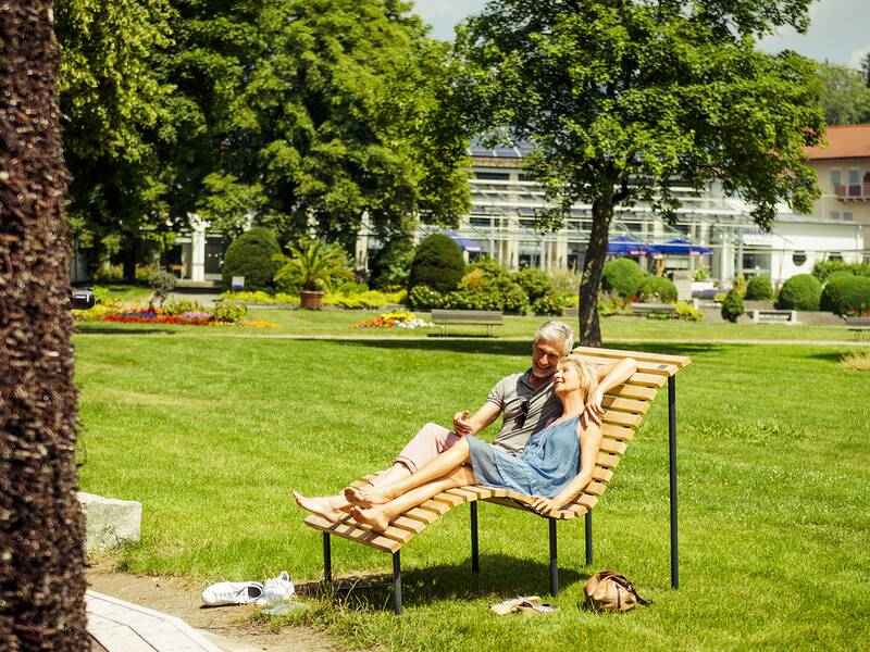 Paar sitzt entspannt auf einer geschwungenen Holzbank im Park, umgeben von grüner Wiese und Bäumen.