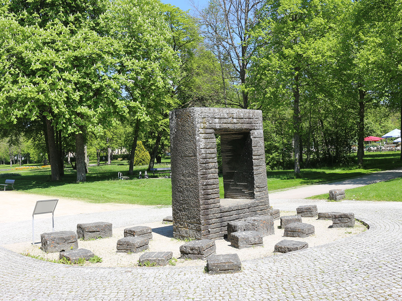 Steinerne quadratische Säule mit rechteckiger Öffnung, umgeben von mehreren kleinen Steinblöcken auf gepflastertem Platz im Park mit Bäumen und Sitzbänken.