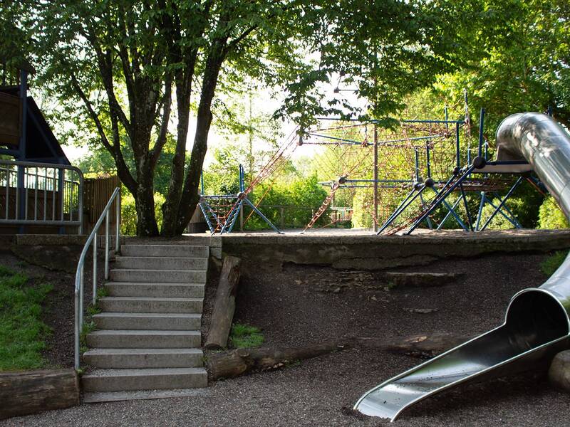 Spielplatz mit Metallrutsche, Klettergerüst und Treppe, umgeben von Bäumen und Grünflächen.