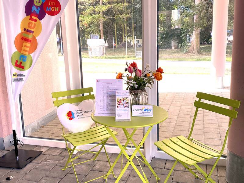 Kleiner runder Tisch mit zwei grünen Klappstühlen vor großen Fenstern, auf dem Tisch ein Blumenstrauß und Informationsmaterialien