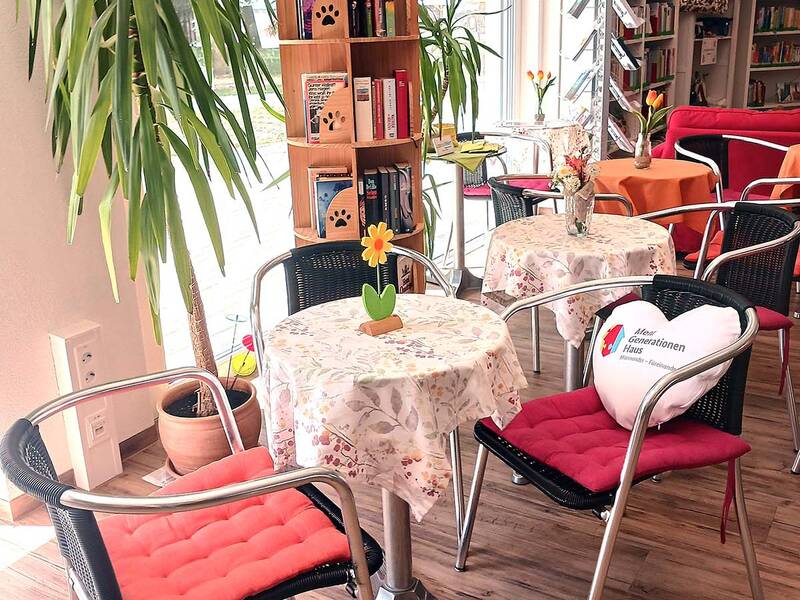 Gemütlicher Cafébereich mit kleinen Tischen, gepolsterten Stühlen, einem Bücherregal und Pflanzen in einem hellen Raum