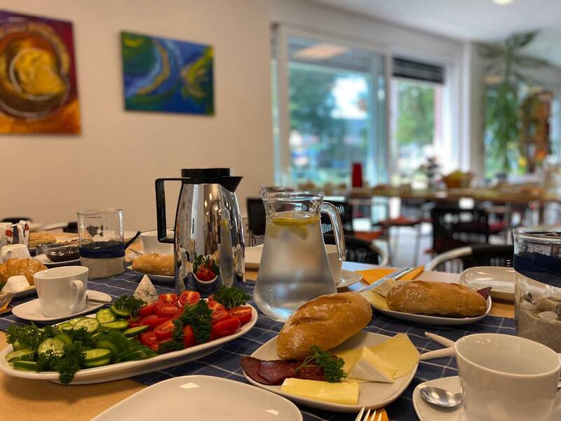 Frühstückstisch mit Brötchen, Käse, Aufschnitt, Gurken, Tomaten, Kaffeekanne und Wasserkaraffe in einem hellen Raum mit großen Fenstern