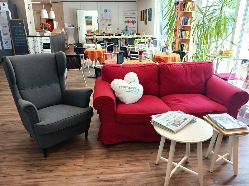 Rotes Sofa mit weißem Herzkissen neben grauem Ohrensessel vor Tischen mit orangenen Tischdecken in hellem Raum