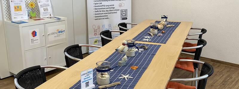 Langer Holztisch mit blauen Läufern und sechs Stühlen mit orangen Sitzkissen in einem Raum mit Informationsständen und bunten Gemälden an der Wand