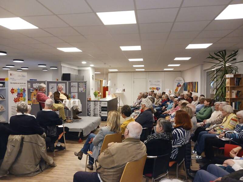 Zuhörende ältere Menschen sitzen in einem Saal und hören zwei Personen auf einer kleinen Bühne zu