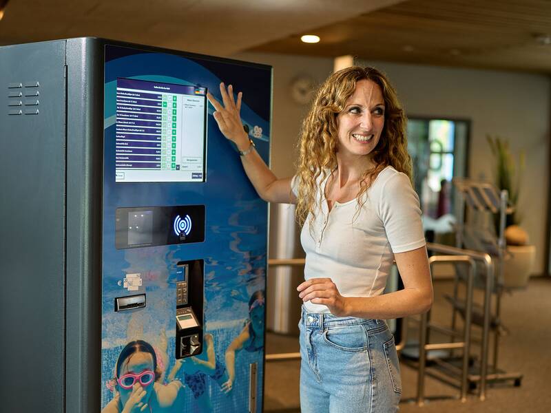 Frau mit lockigem Haar und weißem T-Shirt bedient blauen Kassenautomaten mit Touchscreen und Kartenleser.