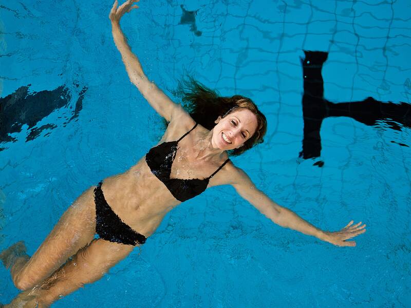 Frau in schwarzem Bikini schwimmt auf dem Rücken in einem Schwimmbecken mit blauer Wasseroberfläche und schwarzen Markierungen.