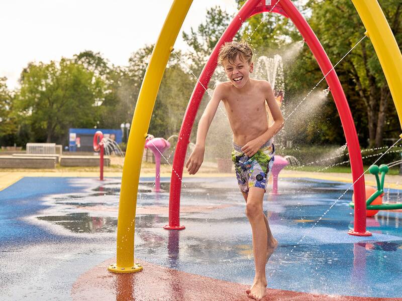 Junge in bunten Badehosen läuft barfuß durch einen Wasserspielplatz mit roten und gelben Bögen, aus denen Wasser spritzt.