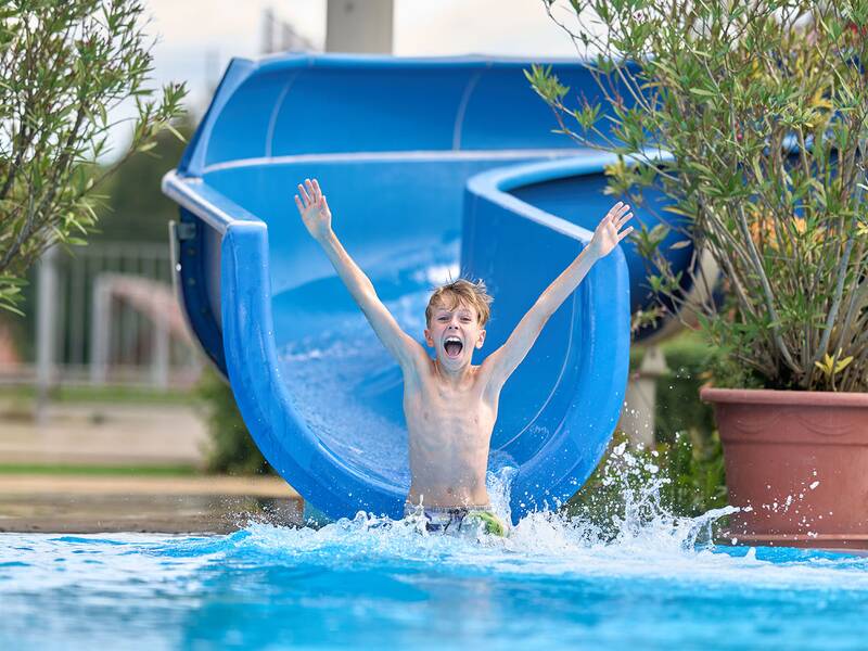 Junge mit erhobenen Armen rutscht aus einer blauen Wasserrutsche in ein Schwimmbecken, umgeben von zwei großen Blumentöpfen mit grünen Pflanzen.