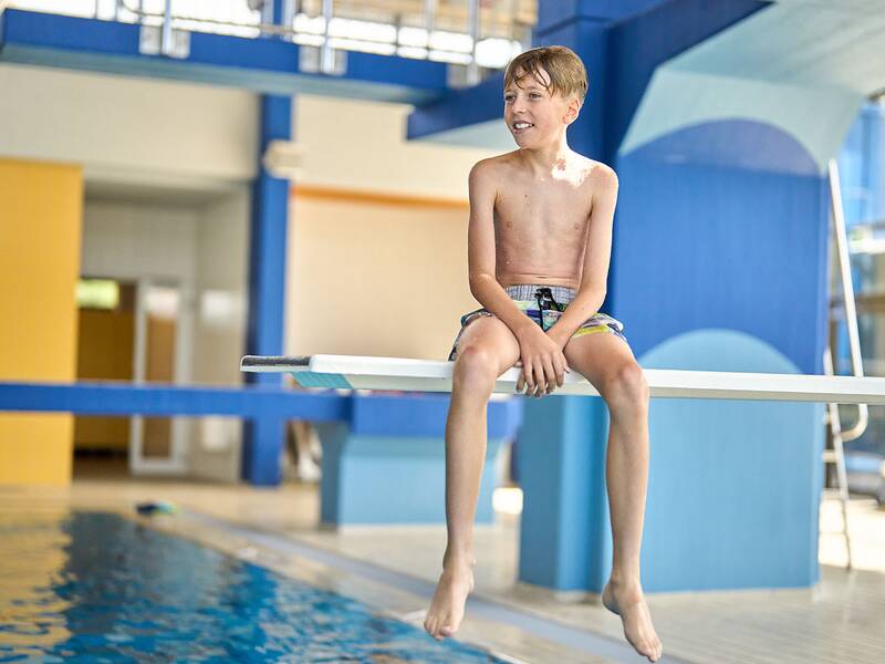 Junge sitzt oben auf einem Sprungbrett in einer Schwimmhalle, trägt Badehose, Beine baumeln über Wasser.