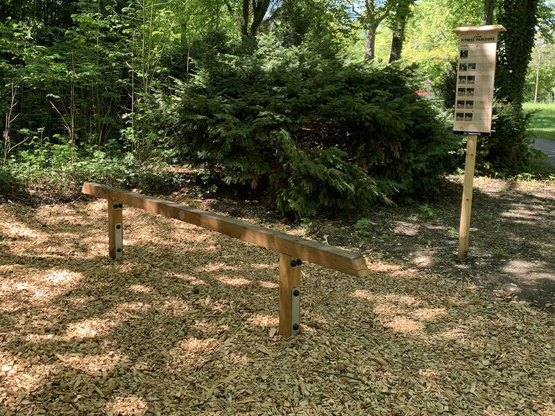 Holz-Balancierbalken auf Holzspänen in einem Park mit Bäumen und einem Informationsschild im Hintergrund.