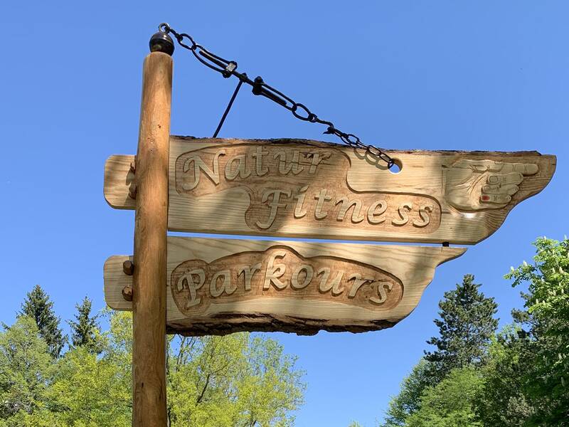 Zwei hölzerne Schilder mit geschnitzter Aufschrift 'Natur Fitness' und 'Parkours' an einem Pfosten vor blauem Himmel und Bäumen.